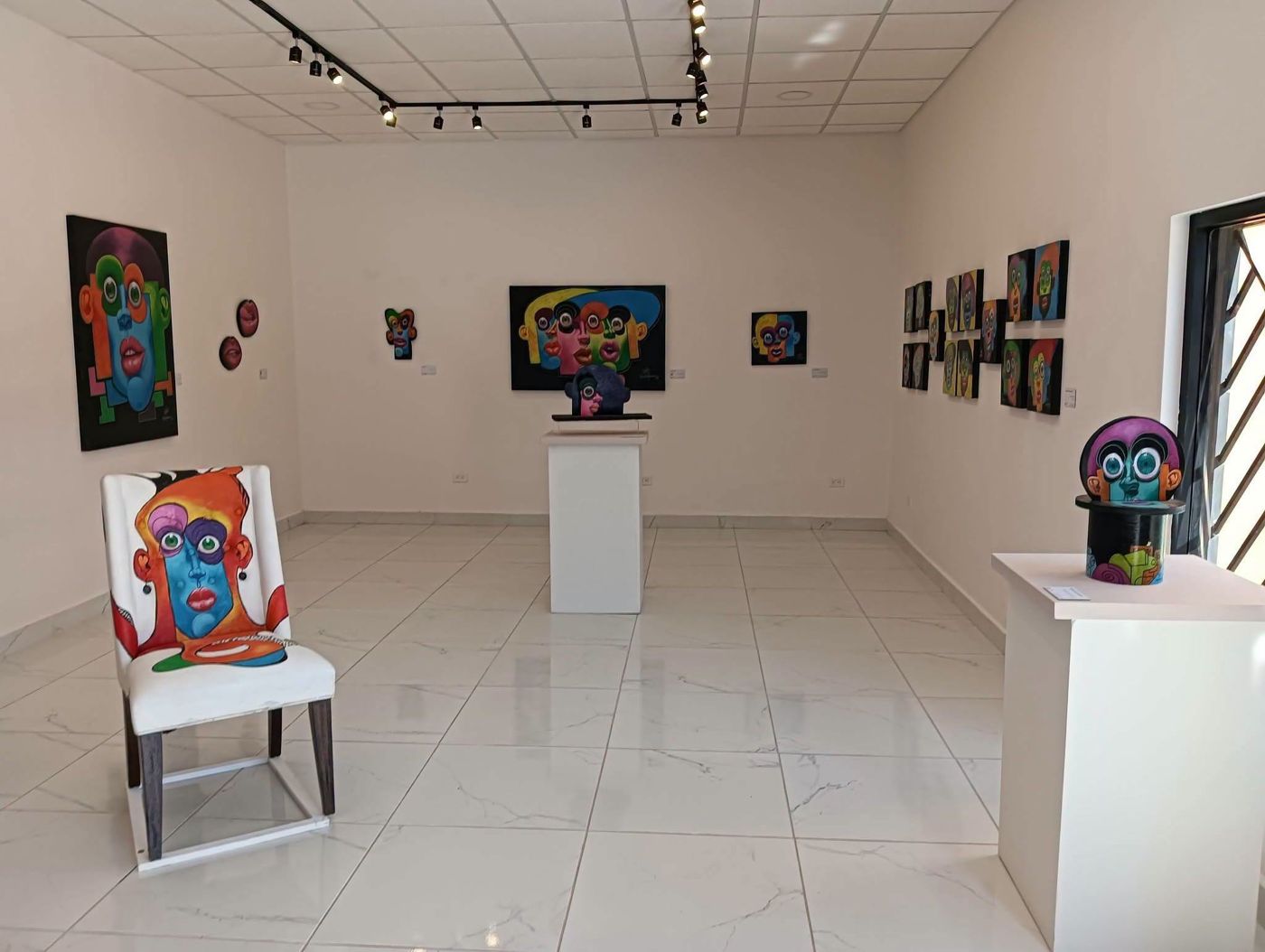 View of the exhibition room with paintings, a painted chair and a sculpture on a pedestal