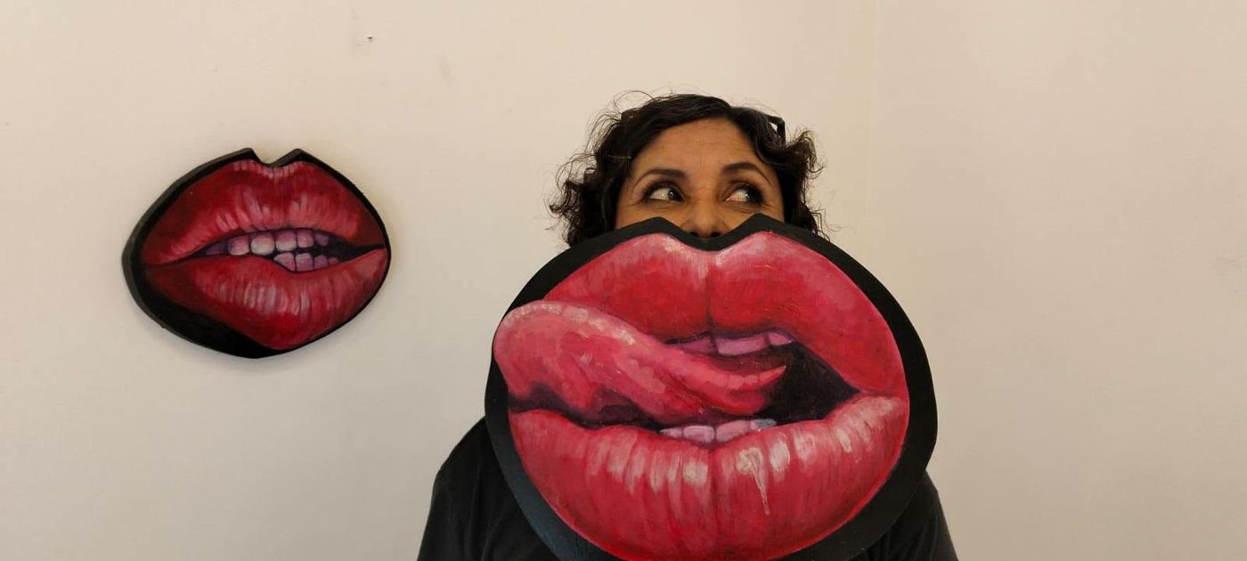 A visitor holding painted lips in front of her mouth, next to the same work on the wall